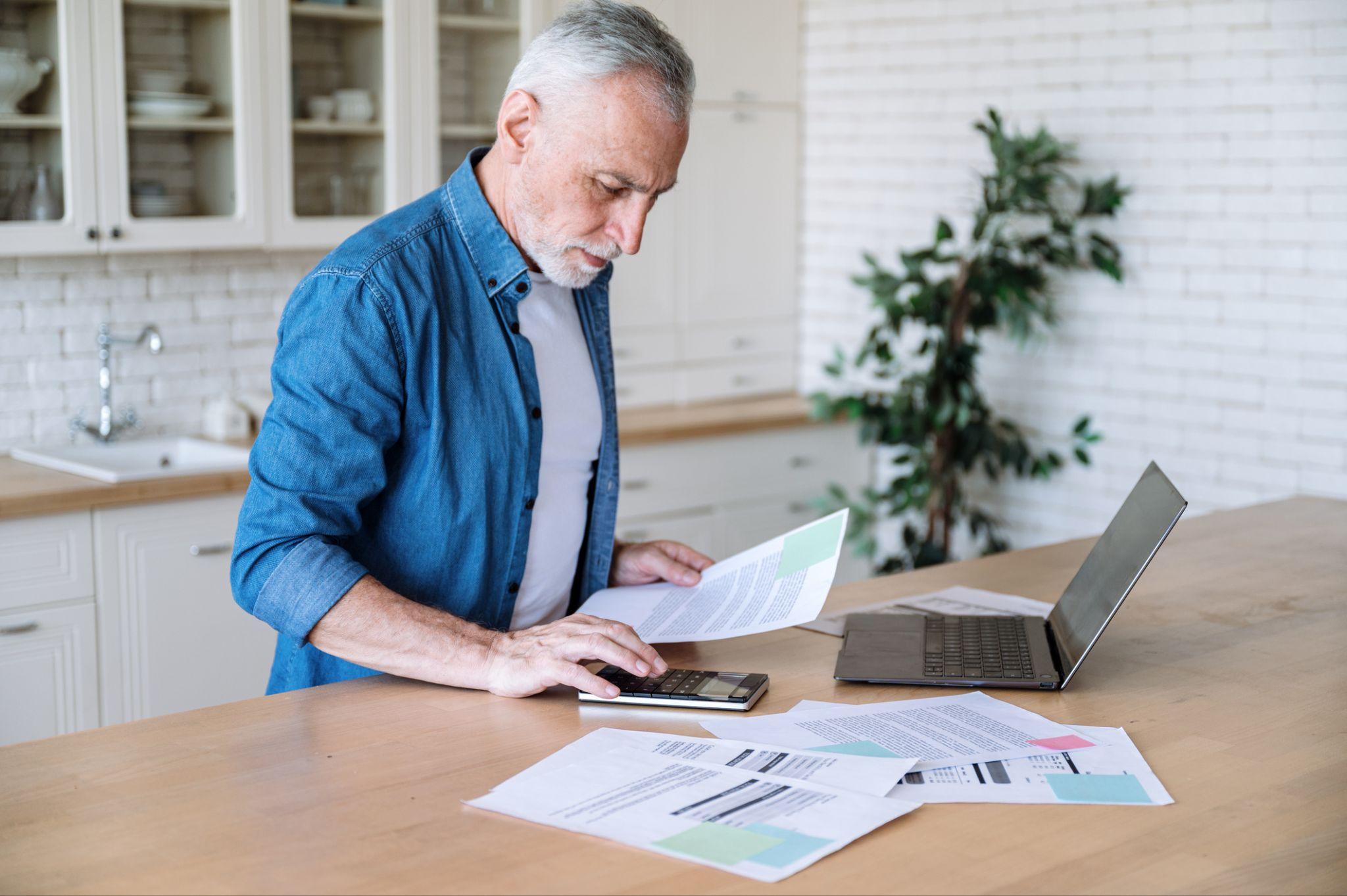 An elderly man in a denim shirt calculating finances with documents and a laptop on the kitchen counter